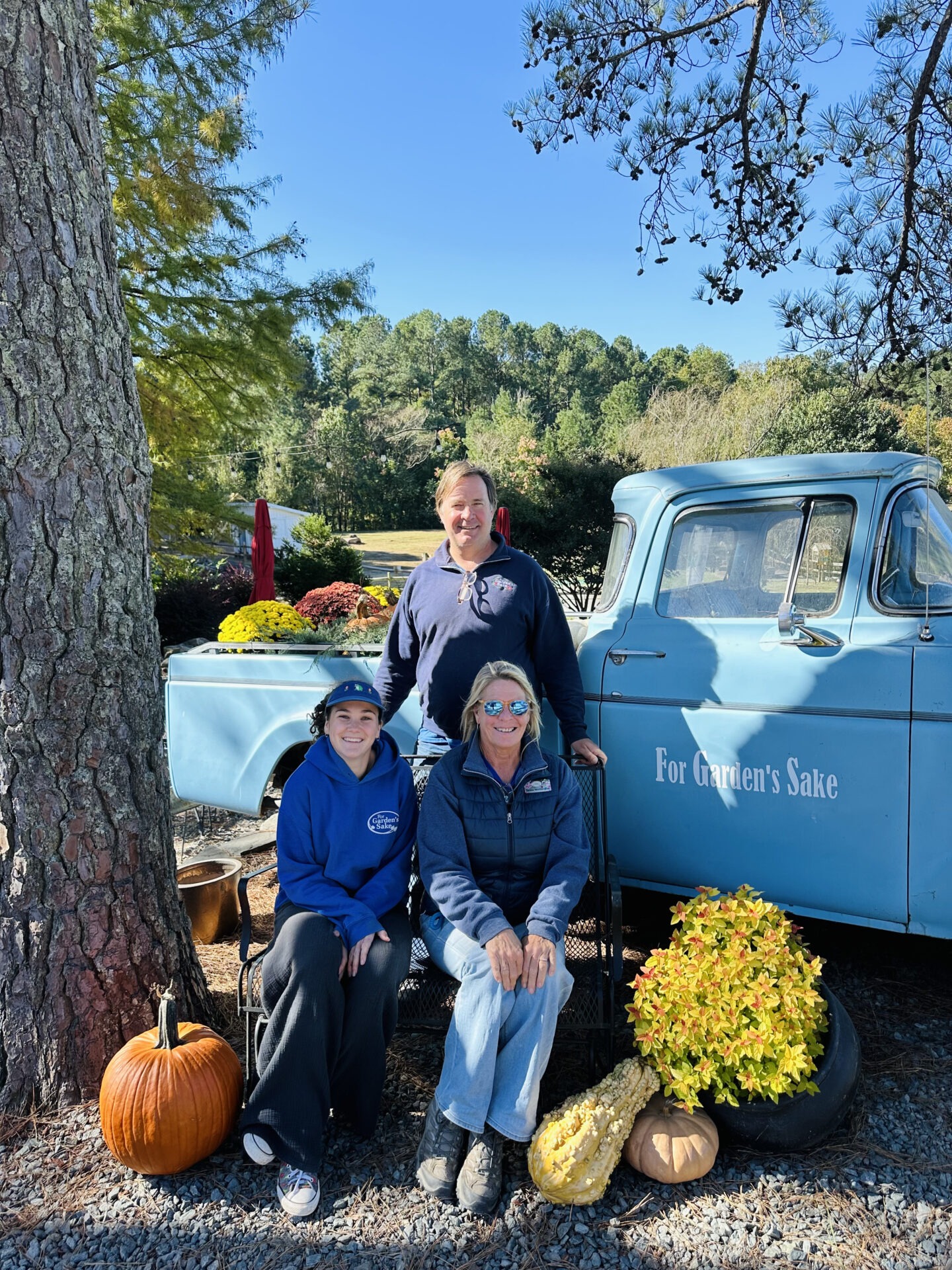 Three people pose with a vintage blue truck surrounded by pumpkins and flowers, in a lush, green outdoor setting under a clear sky.