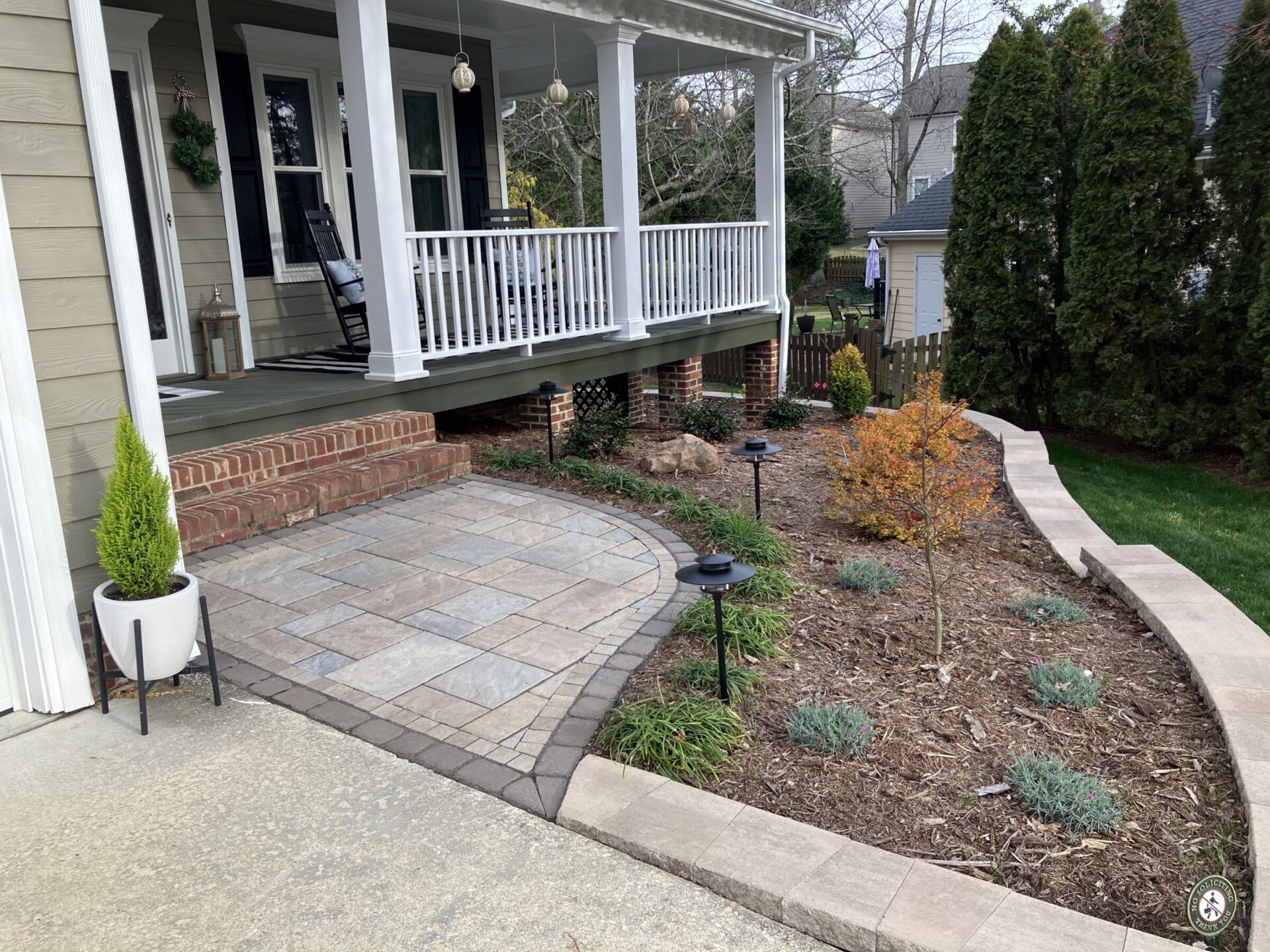 A house with a spacious front porch, brick foundation, landscaped garden, small trees, and tidy walkway. Calm residential setting with a modern, welcoming feel.