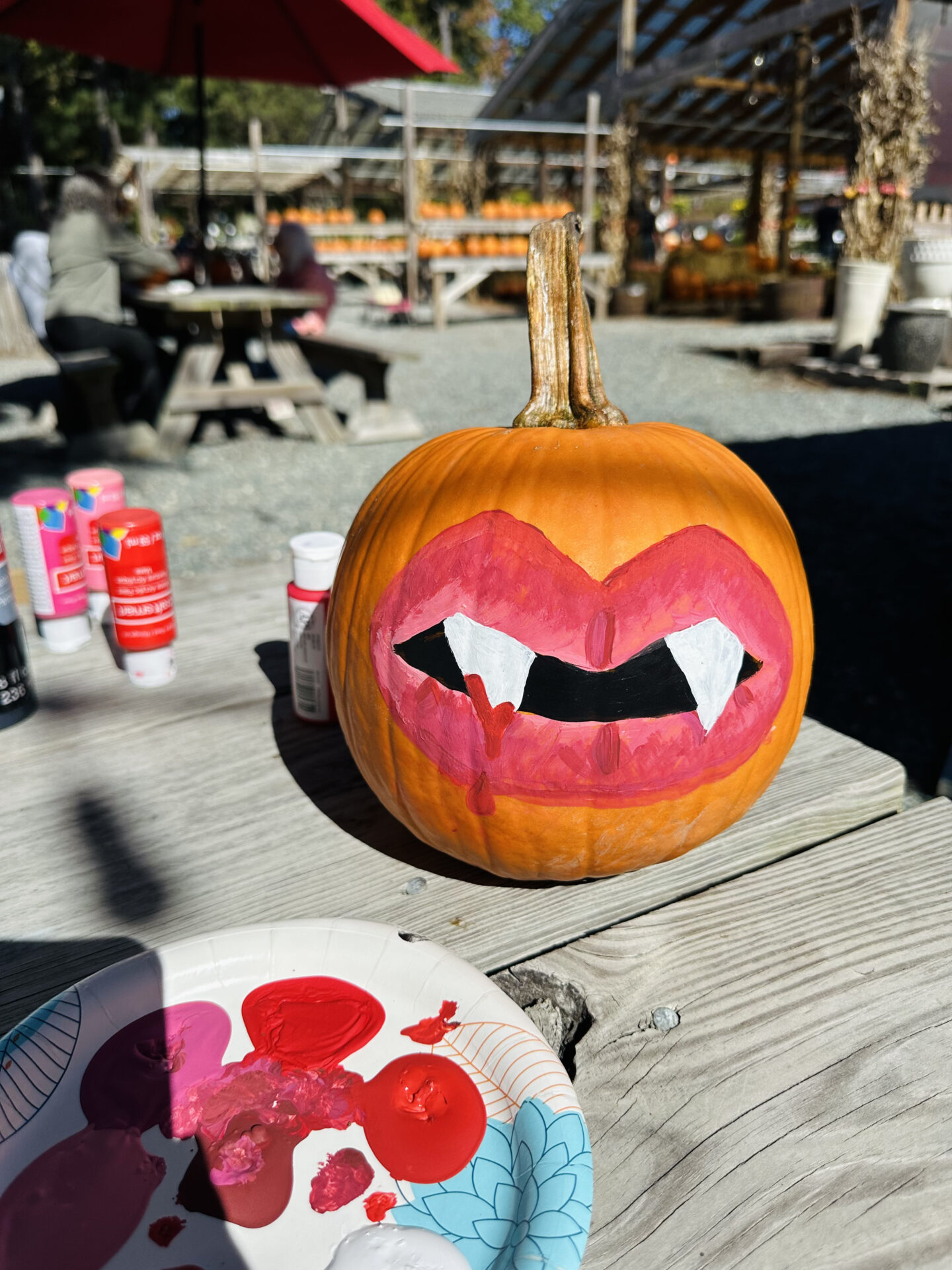 A pumpkin painted with vampire lips and fangs sits on a picnic table, surrounded by paint supplies and outdoor seating in bright sunlight.