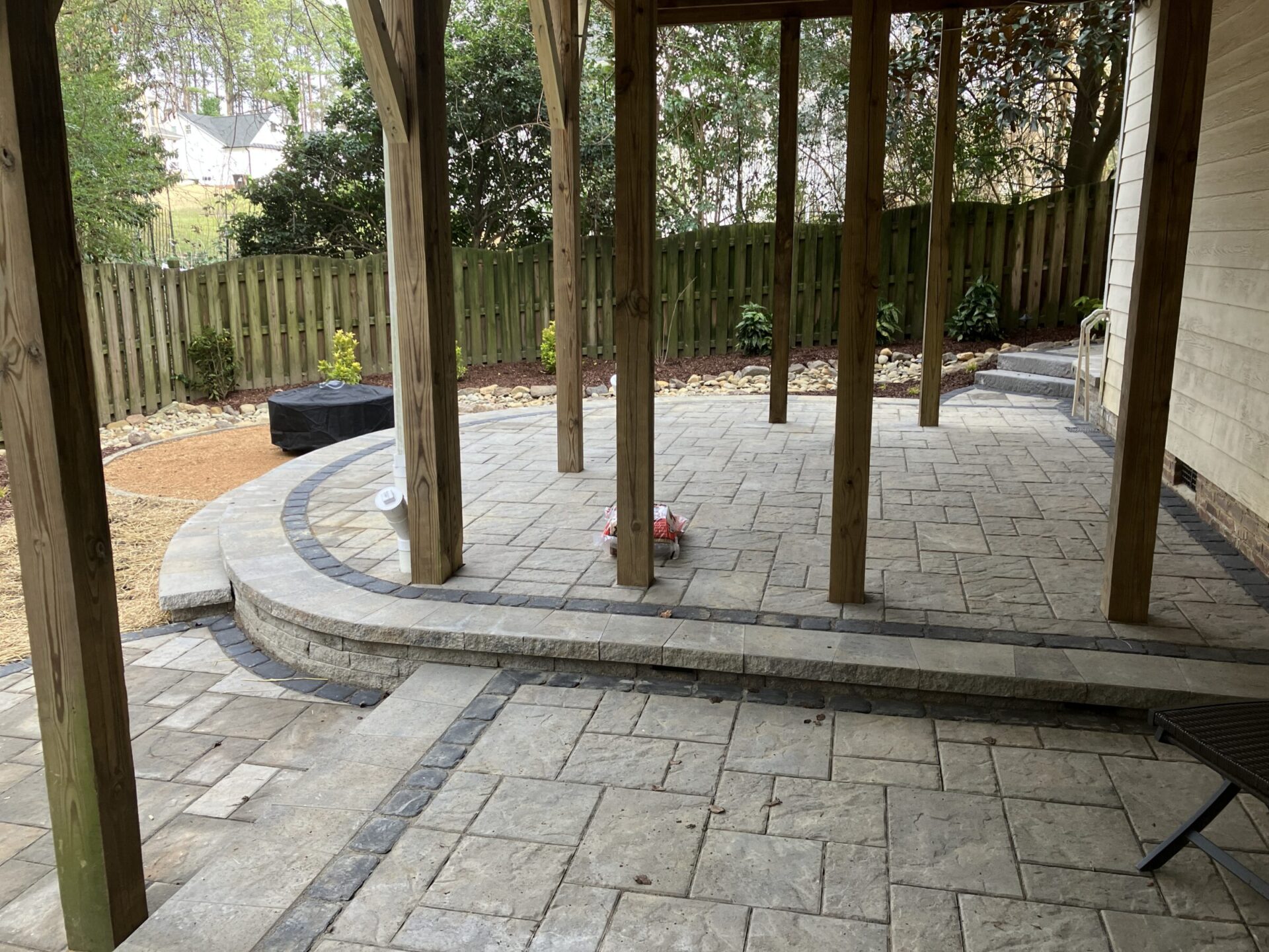 A backyard patio features stone paving under a wooden pergola. The space is bordered by a wooden fence and landscaped with small plants.