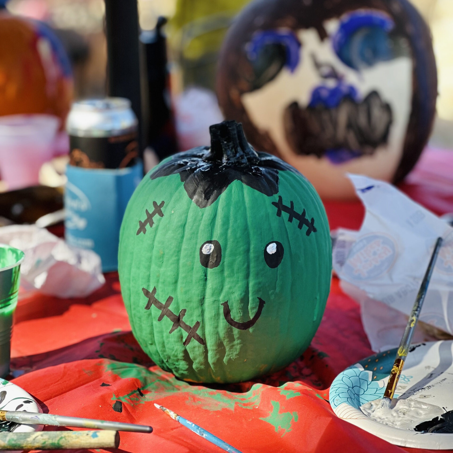 Green Frankenstein-themed pumpkin with smiling face, surrounded by art supplies and drinks on a red table. Background shows another painted pumpkin.