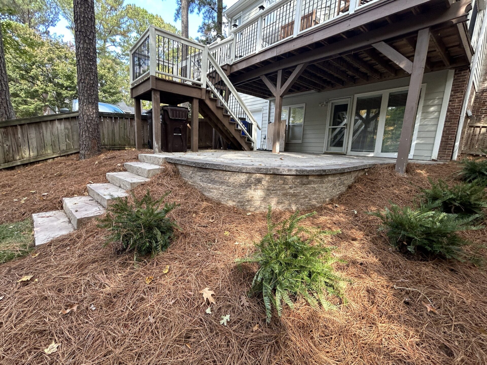 A wooden deck with stairs overlooks a landscaped yard. Pine mulch covers the ground, surrounded by small green shrubs and tall trees.