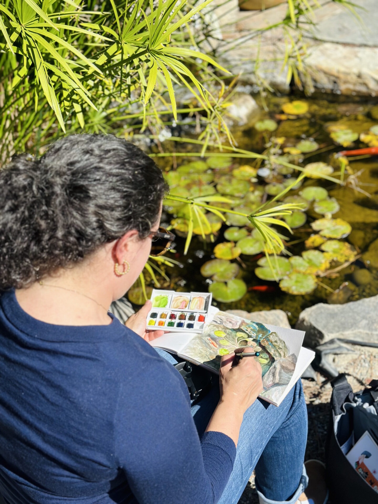 A person sitting by a pond, painting with watercolors, surrounded by lush plants and lily pads, enjoying a serene outdoor setting.