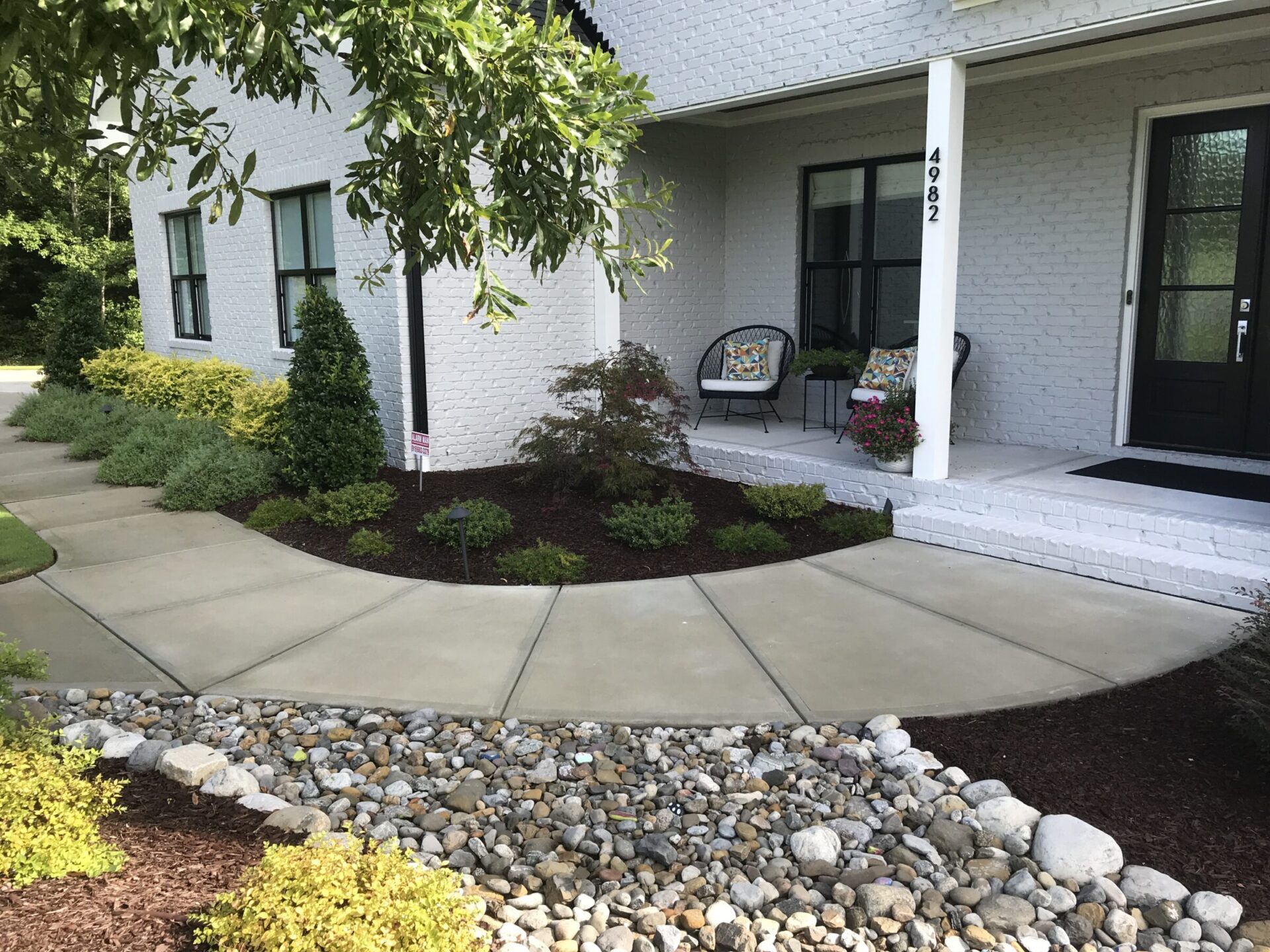 A white brick house features a landscaped front yard with a concrete path, decorative chairs, and a small porch surrounded by greenery and stones.