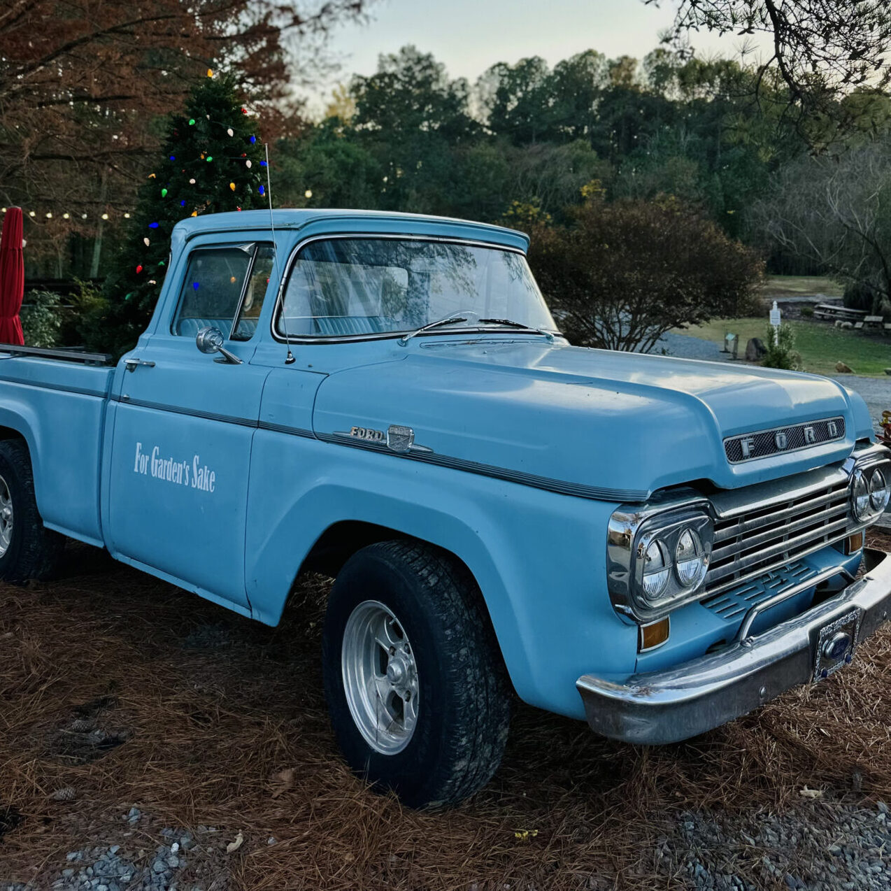 A vintage blue Ford truck is parked outdoors next to a decorated Christmas tree, surrounded by autumn trees and a peaceful rural landscape.