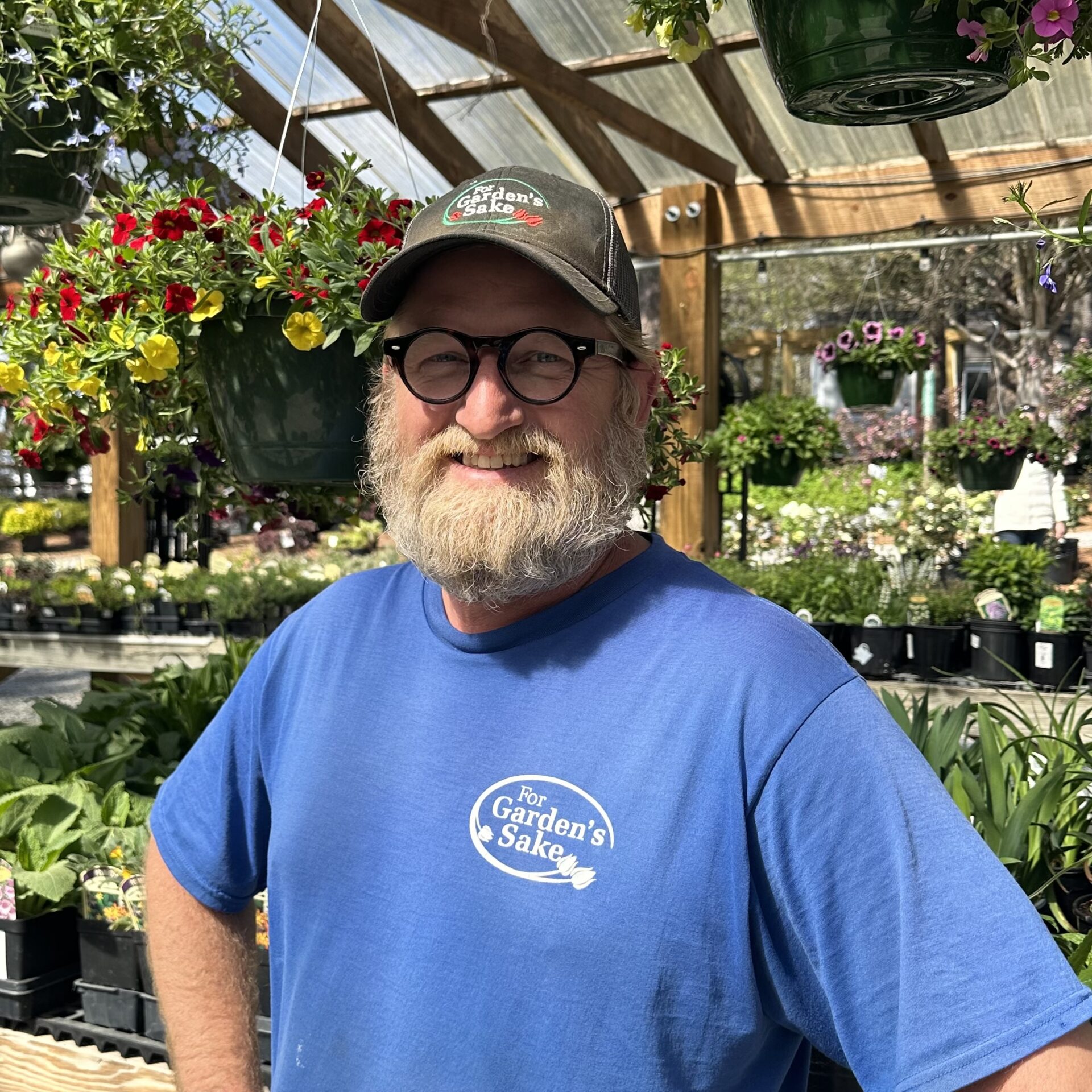 A person in a greenhouse, wearing glasses and a cap, stands among colorful hanging flowers and potted plants, smiling warmly at the camera.