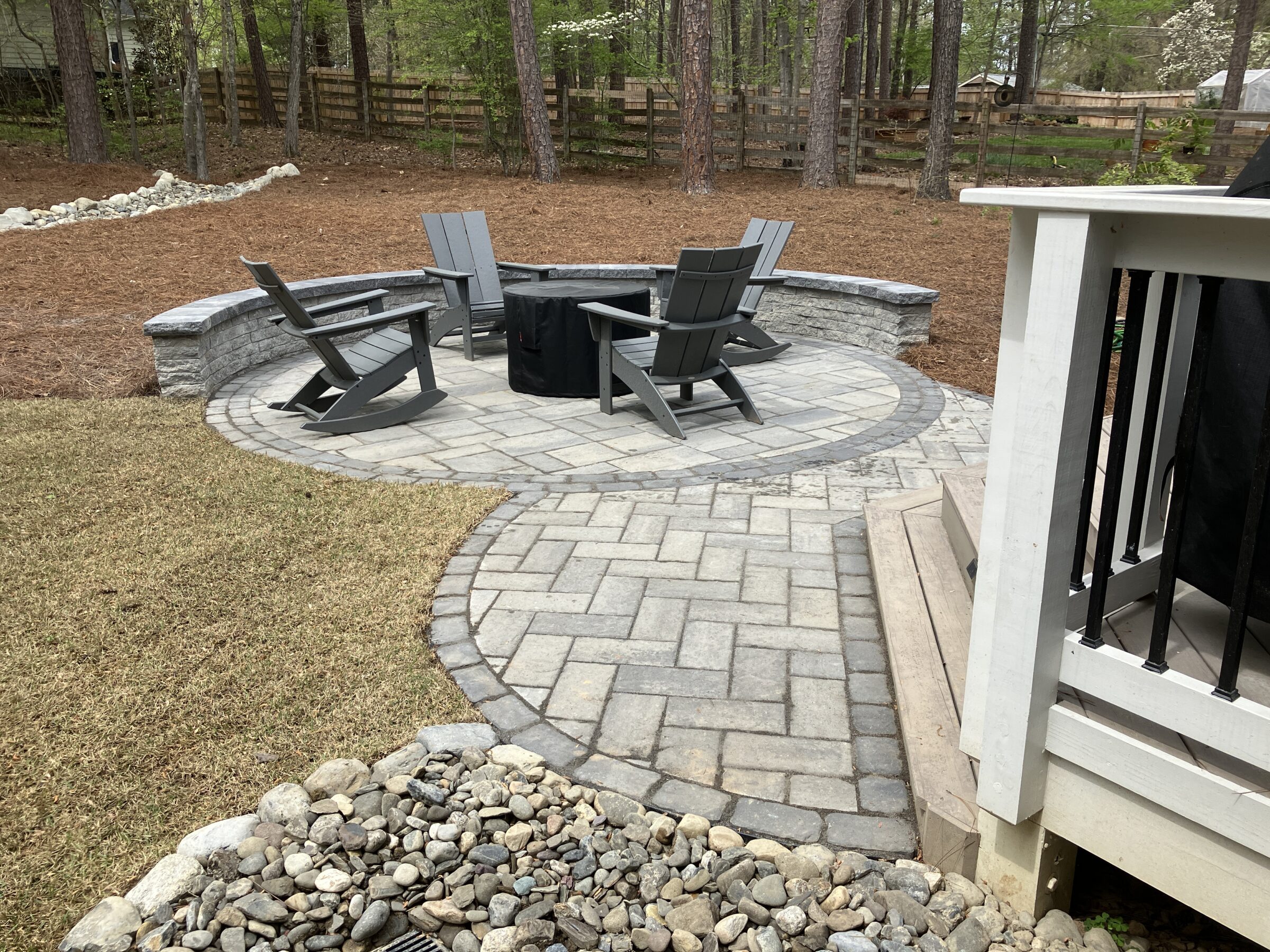 A stone patio with Adirondack chairs encircling a fire pit, surrounded by trees and manicured lawn in a backyard setting.