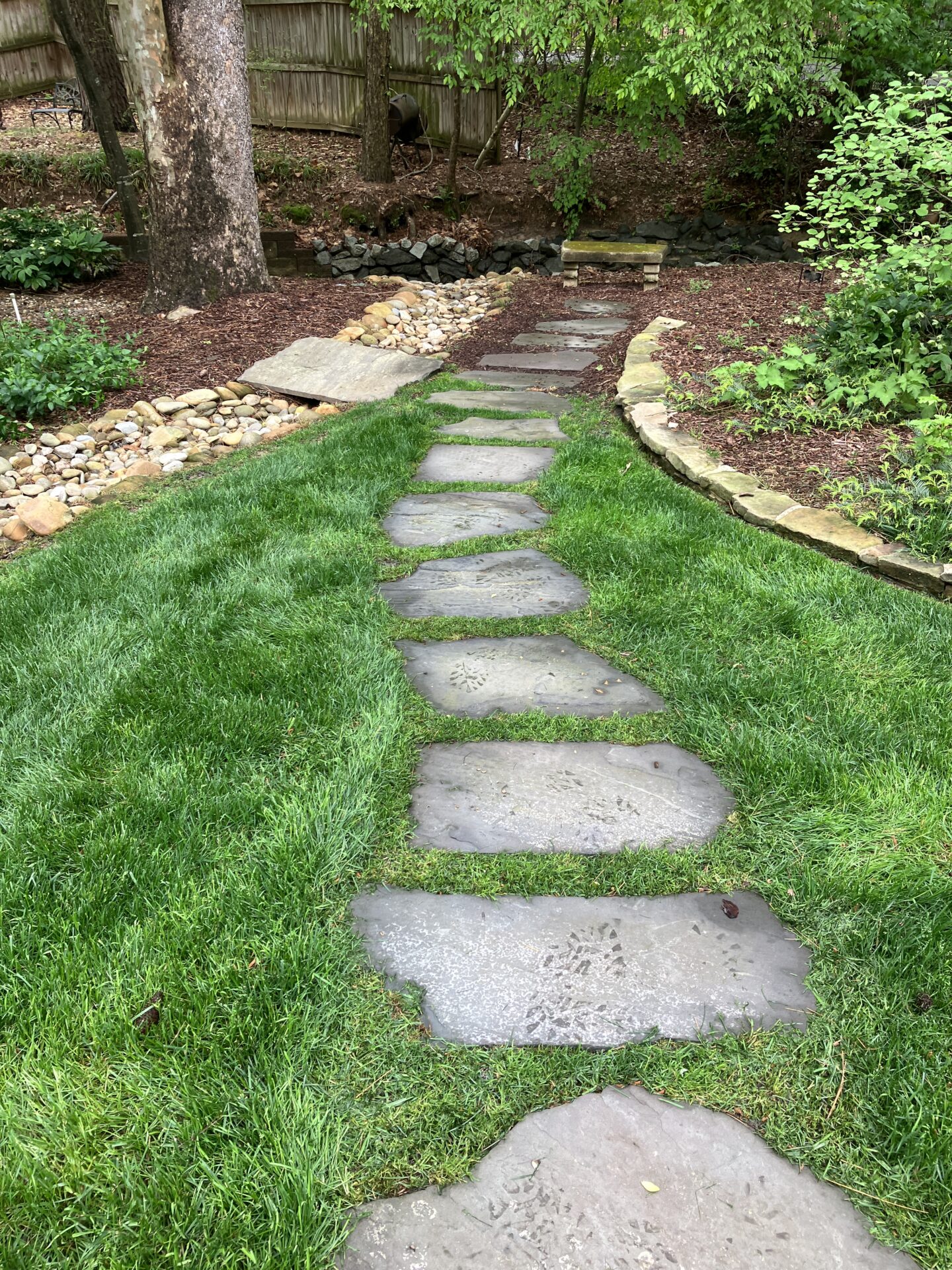 A stone pathway meanders through a lush garden, bordered by trees, rocks, and mulch, creating a serene, natural environment under a cloudy sky.