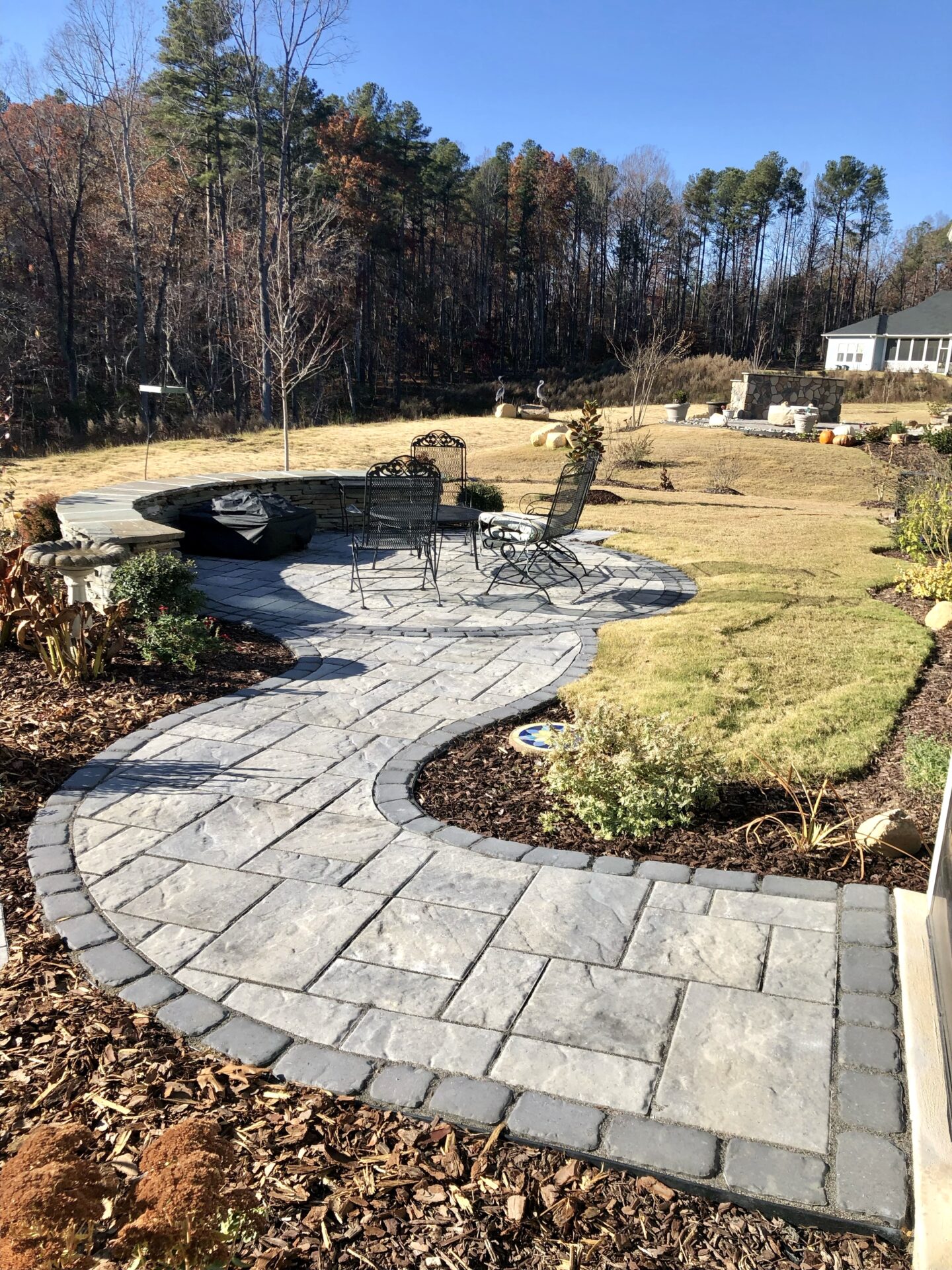 Curved stone path leads to a patio with chairs, surrounded by landscaped garden and trees under a clear sky.