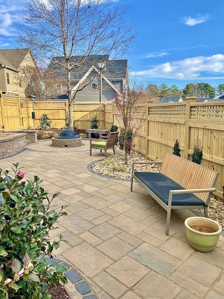 A modern backyard features a paved patio, wooden fencing, seating, a fire pit, potted plants, and a clear blue sky overhead.