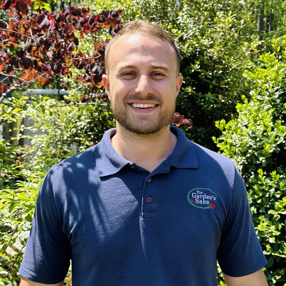 A person stands outdoors smiling, wearing a navy blue polo shirt with a logo. The background features lush green foliage and sunlight.