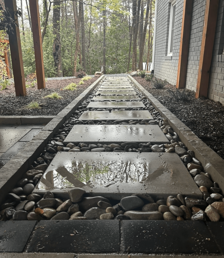 Stone pathway with stepping stones and pebbles, flanked by a house and garden. Wet surface reflects surrounding trees and overcast sky.