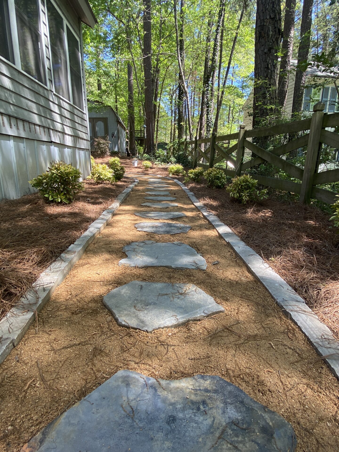 Stone path lined with plants leads through a wooded area, adjacent to a wooden fence and a building with a paneled exterior.