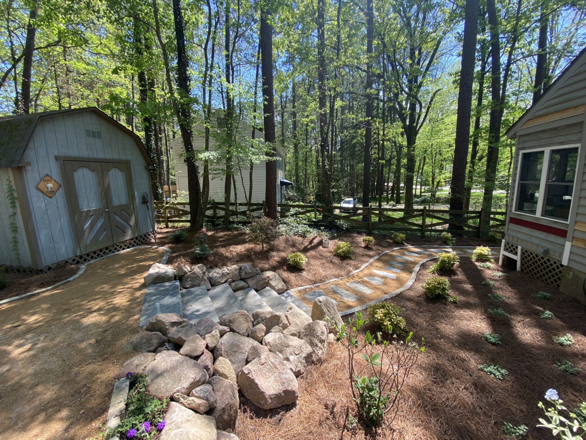 A serene backyard garden with a stone path, surrounded by trees and bordered by a small wooden shed and house, under sunlight.