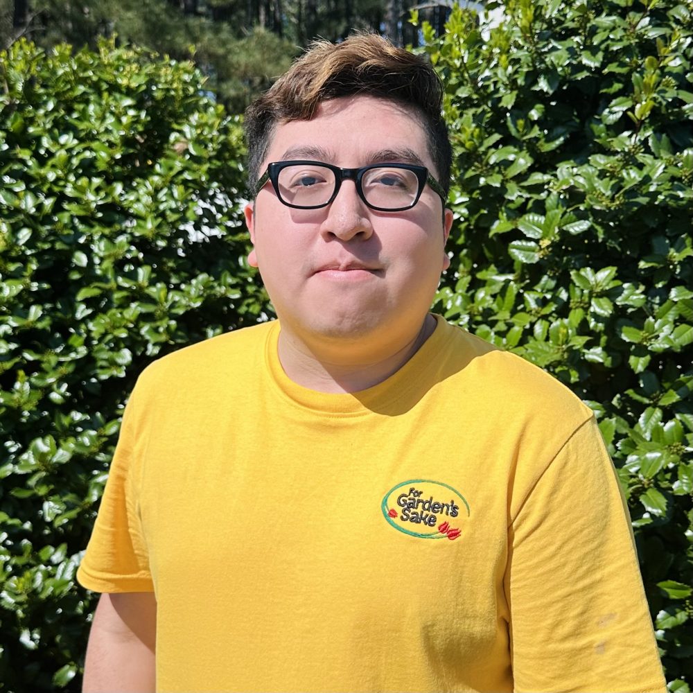 A person with glasses, wearing a yellow shirt, stands in front of lush green foliage under a sunny sky.