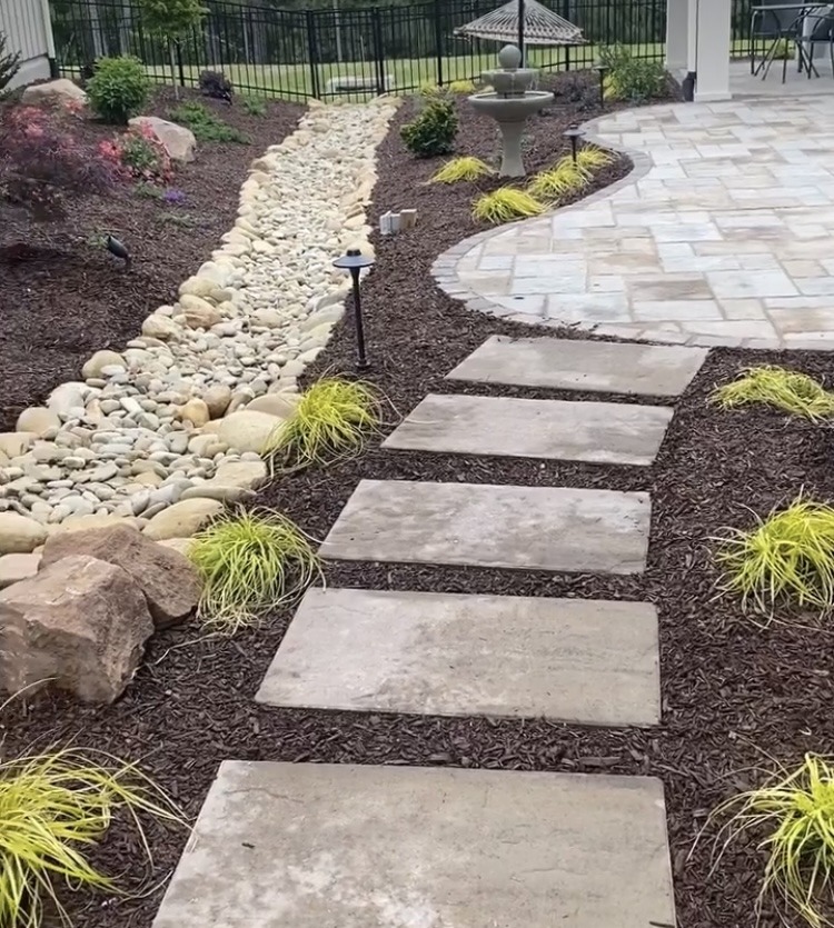 A landscaped garden features a stone path, dry creek bed, patio, small fountain, and decorative plants, enclosed by a black metal fence.