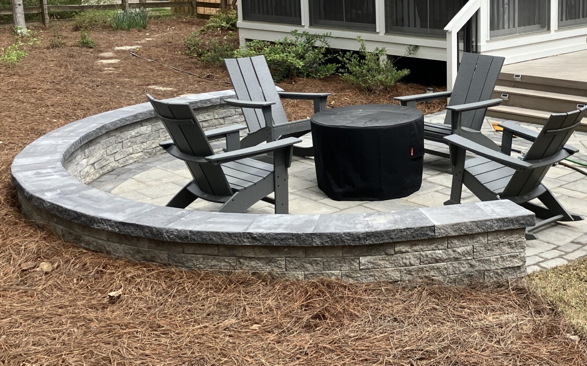 A paved patio features Adirondack chairs around a covered fire pit, surrounded by a low stone wall, with mulch and plants nearby.