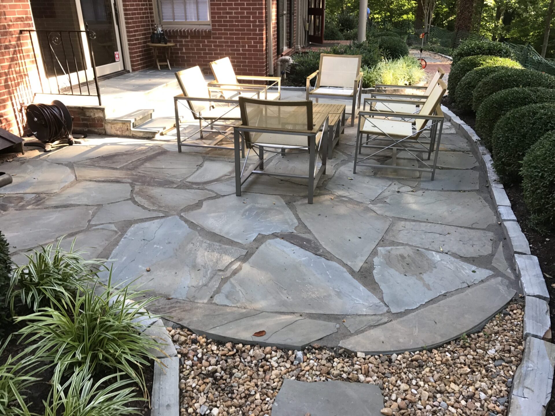 A stone patio with chairs and a table, surrounded by greenery and a brick wall. Peaceful outdoor setting with natural elements.