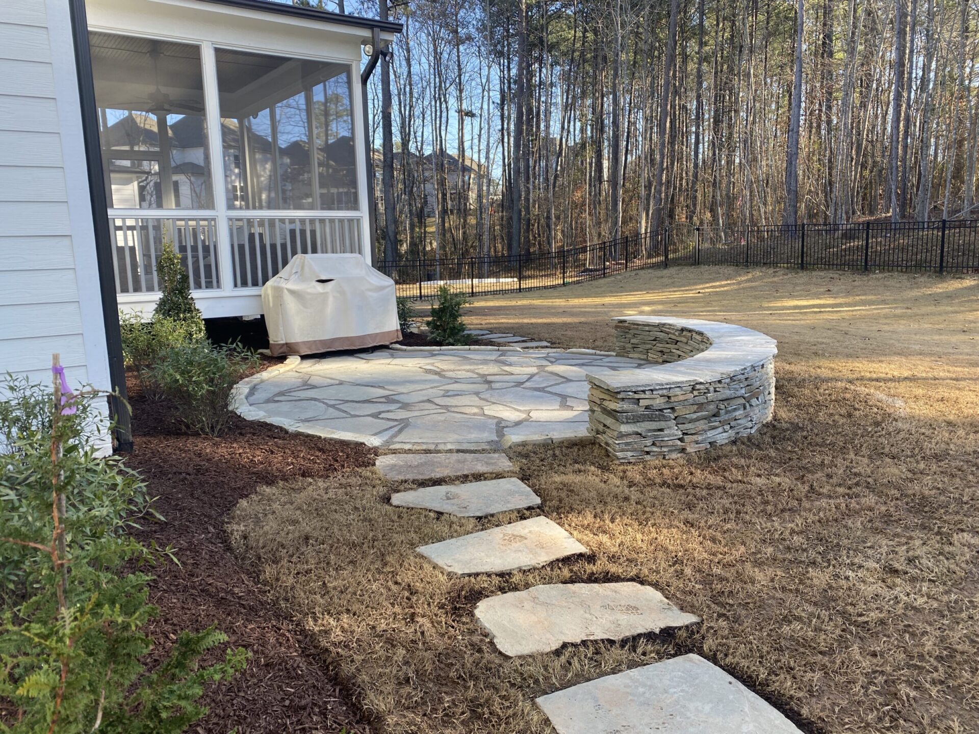 A backyard features a stone patio with a curved stone wall and stepping stones, surrounded by trees and bordered by a white porch.