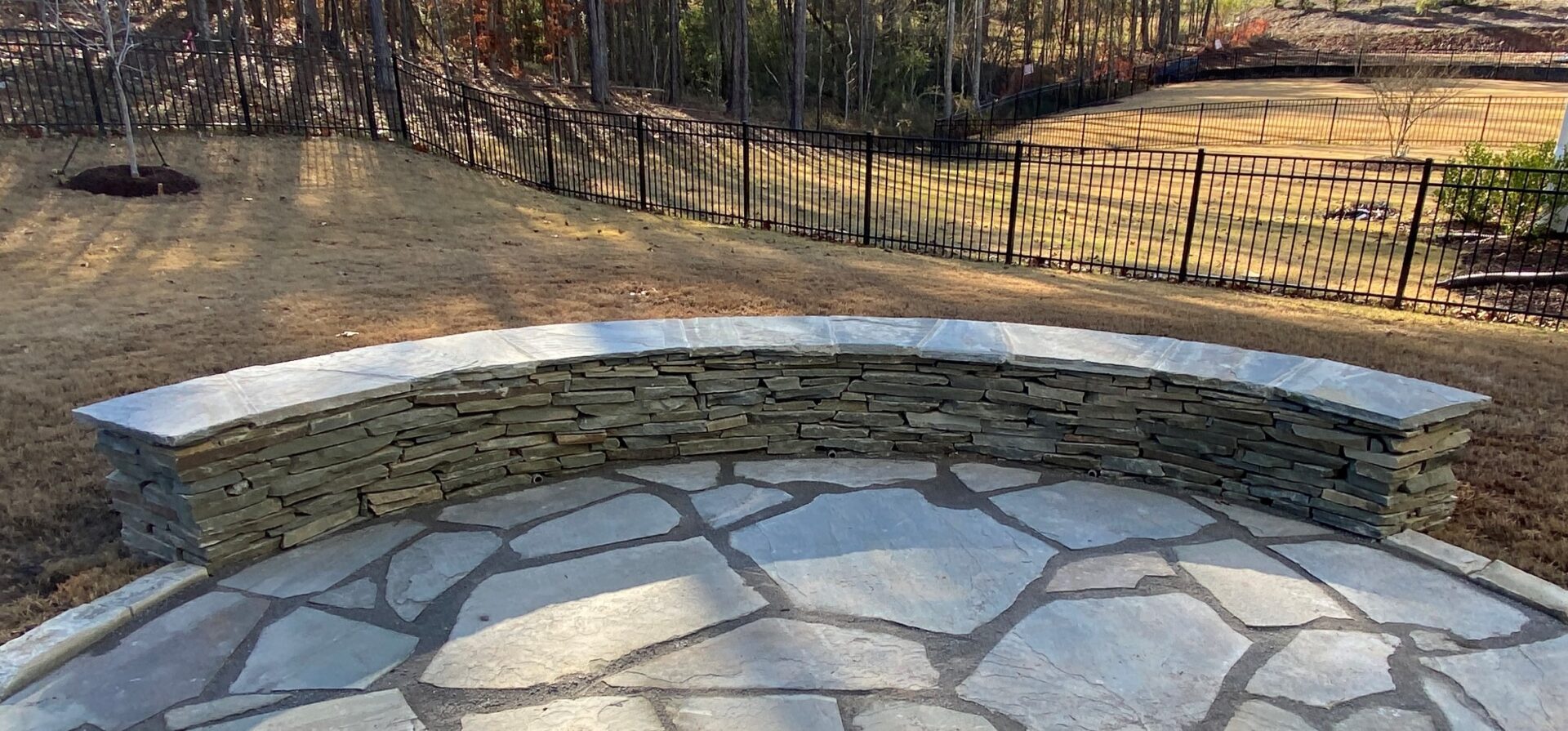 Stone patio with curved stone bench, surrounded by a grassy yard and black fence, bordered by wooded area in the background.