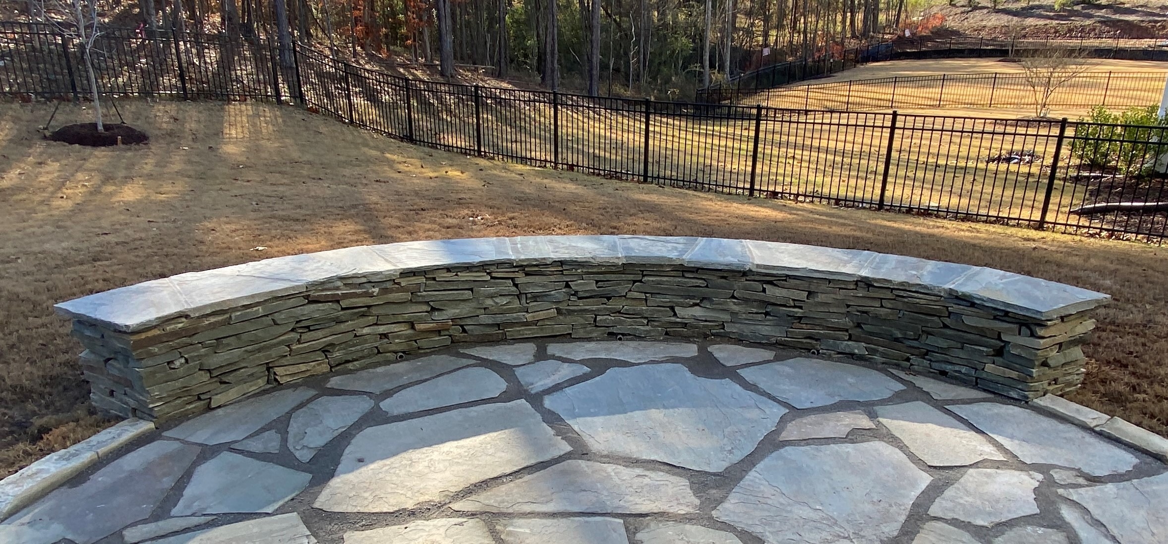 Stone patio with curved stone bench, surrounded by a grassy yard and black fence, bordered by wooded area in the background.