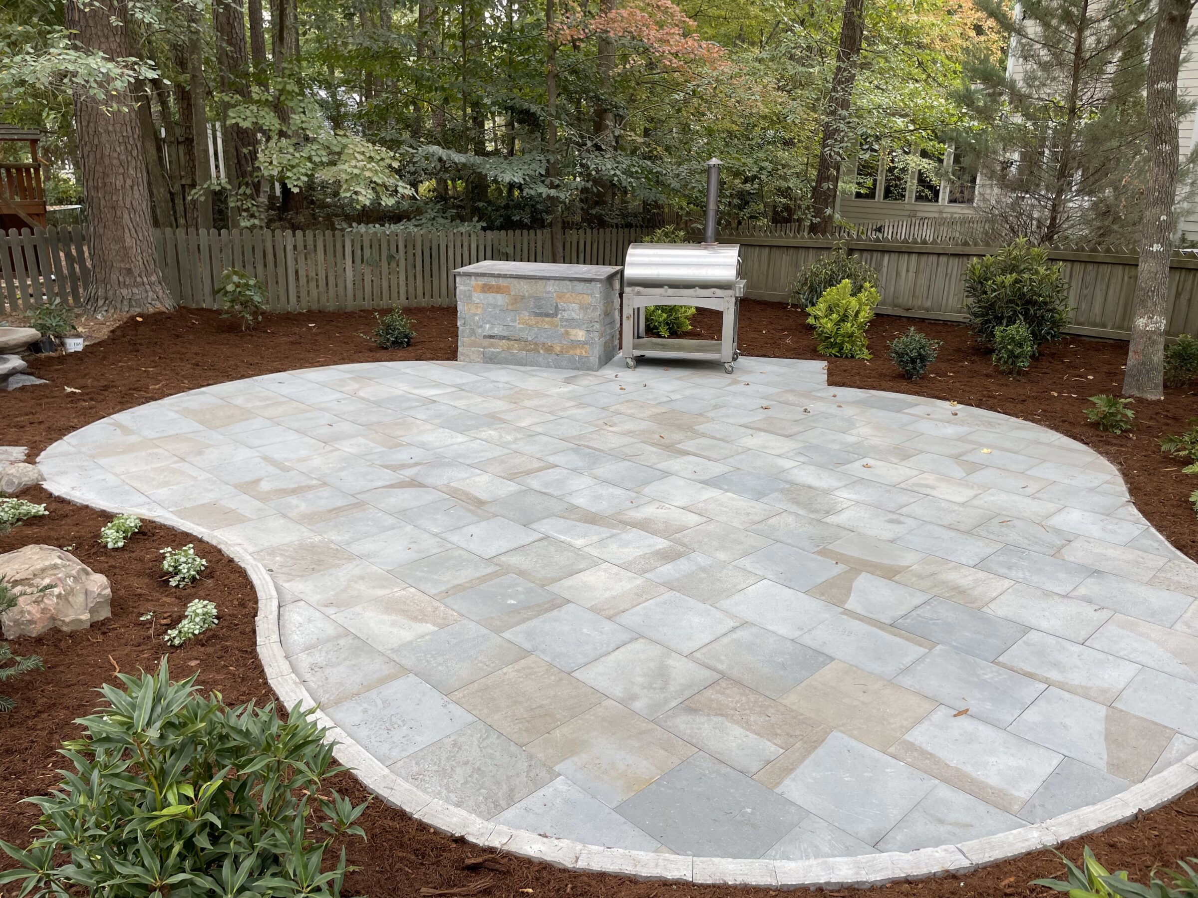 A stone patio with grill and counter in a fenced backyard, surrounded by trees and shrubs, providing a peaceful outdoor setting.