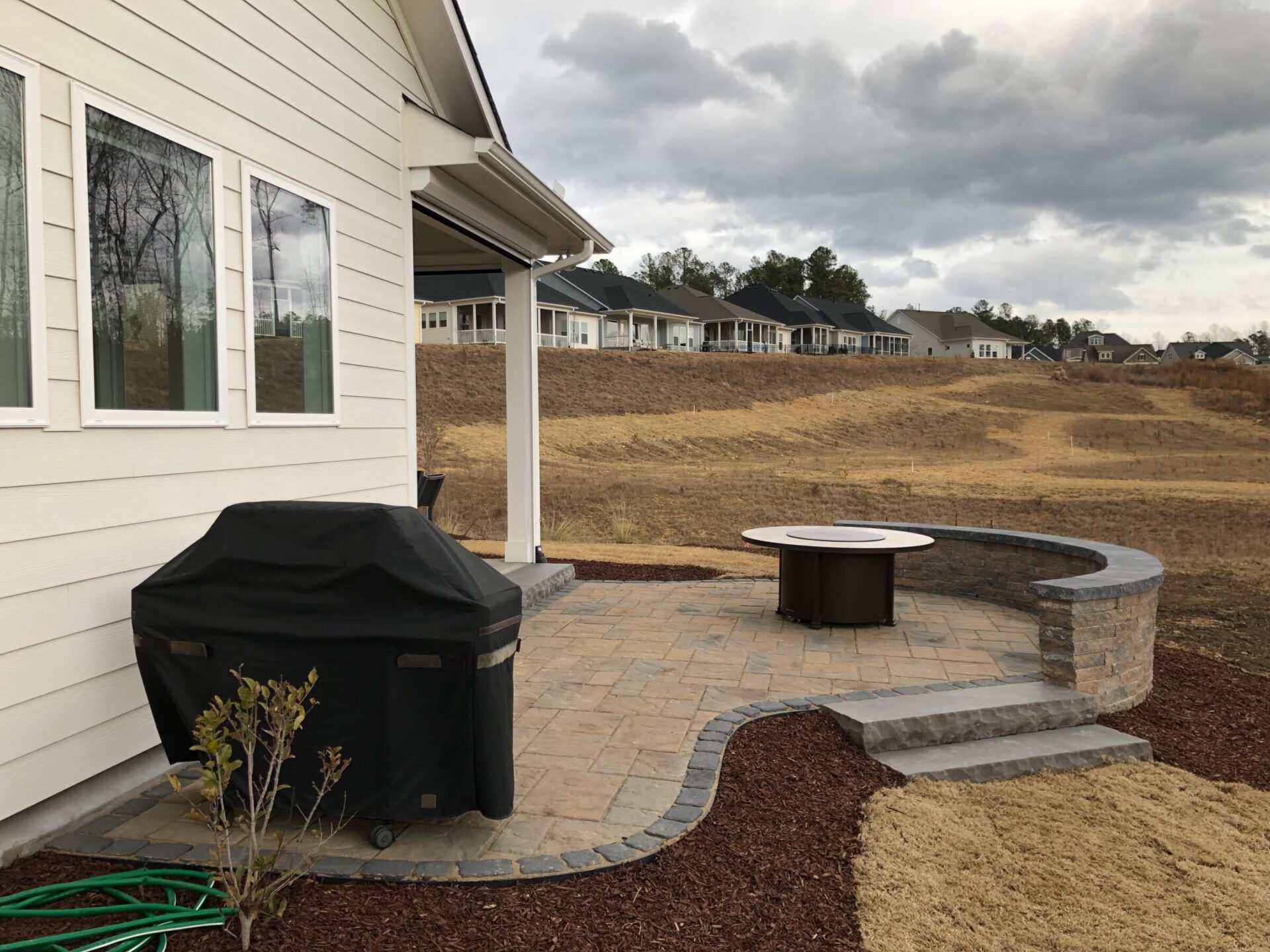 A backyard patio with a grill and circular fire pit, surrounded by bare land and rows of houses under a cloudy sky.