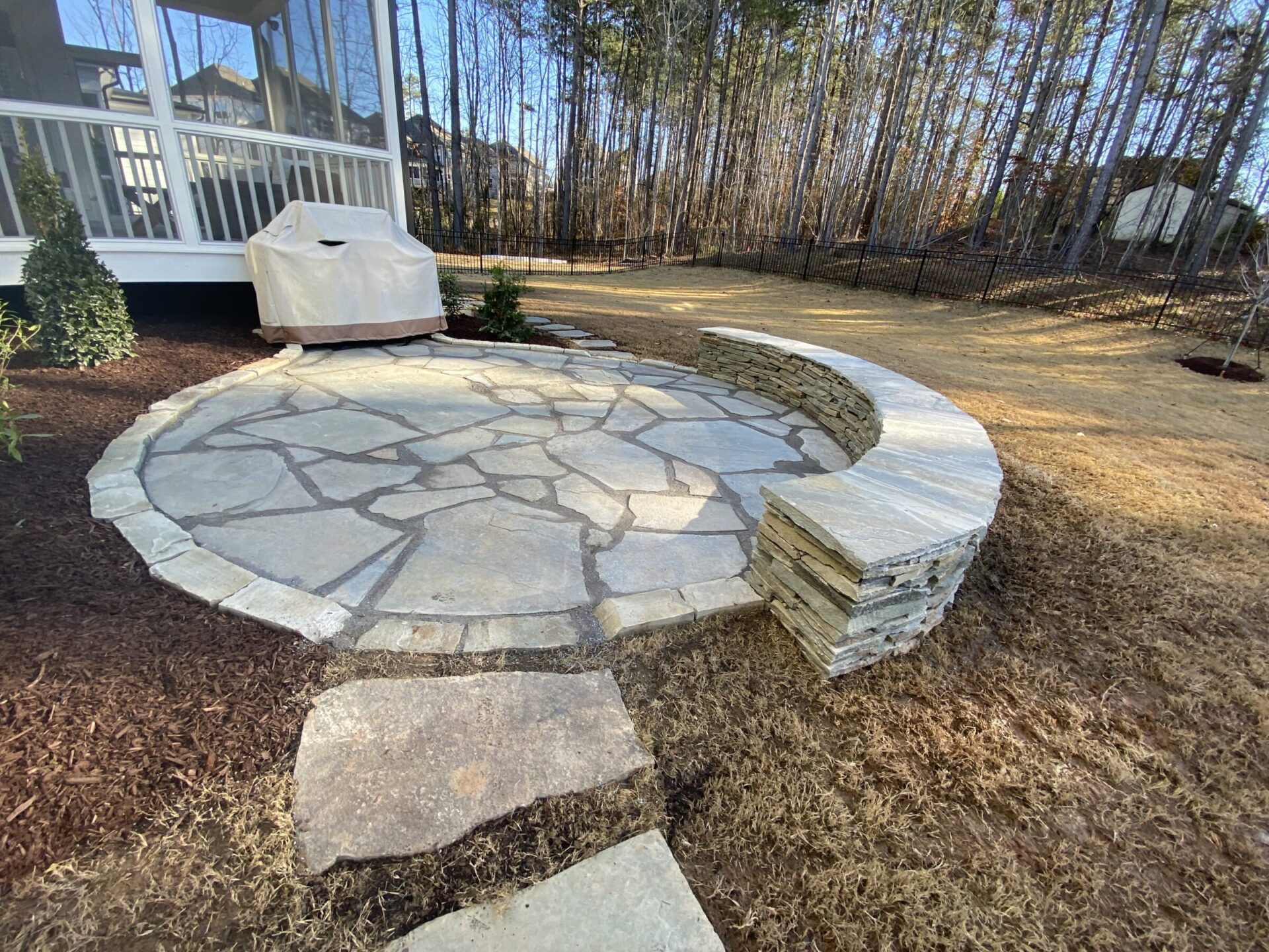 Circular stone patio with curved stone bench, surrounded by mulch and grass. Trees line the background beside a covered porch.