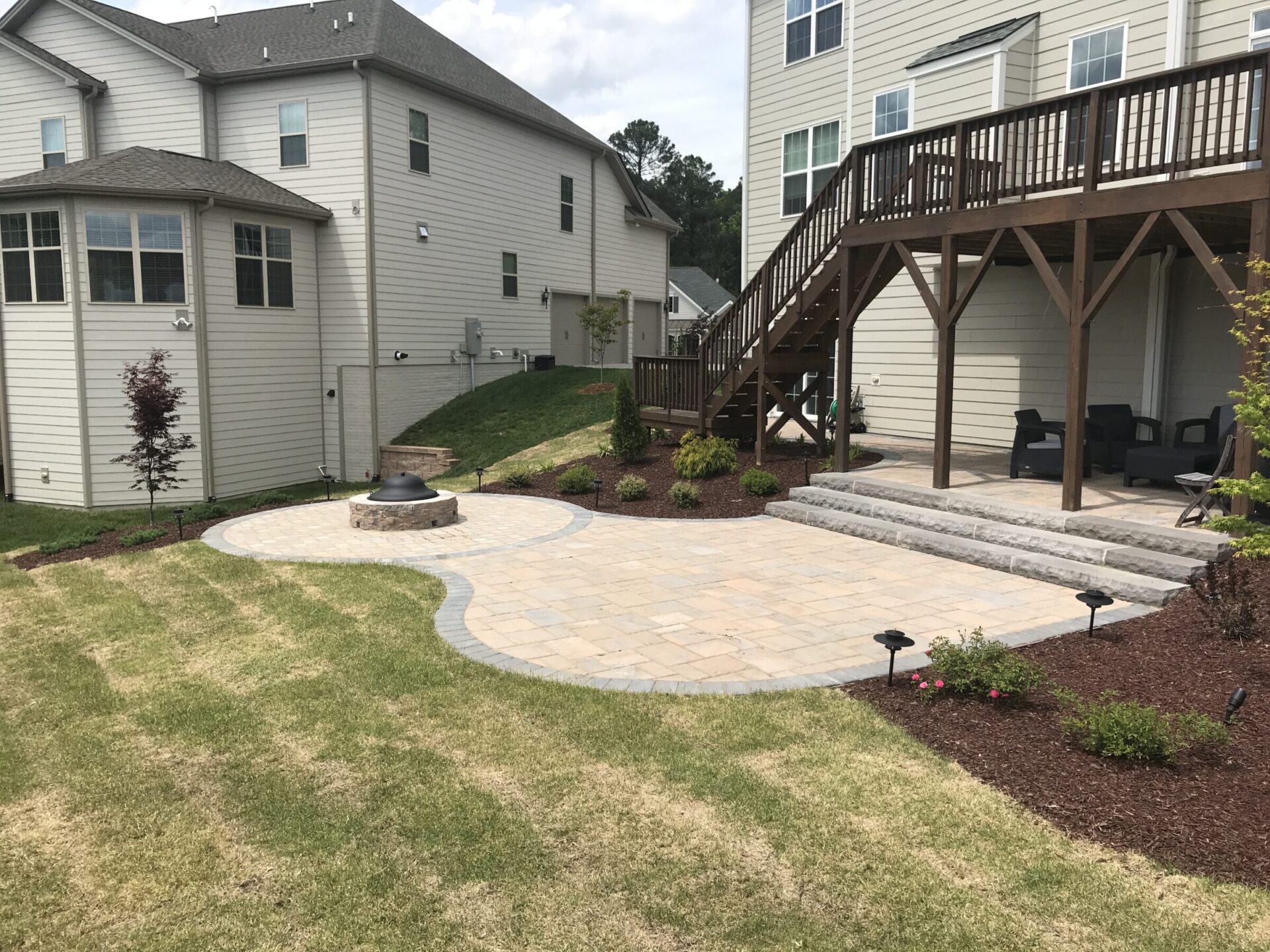 A landscaped backyard features a patio with a fire pit, surrounded by neatly trimmed grass and two-story houses with wooden decks.