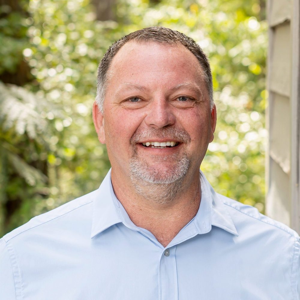 A person in a light blue shirt smiles outdoors, surrounded by lush green foliage and sunlight filtering through the trees in the background.