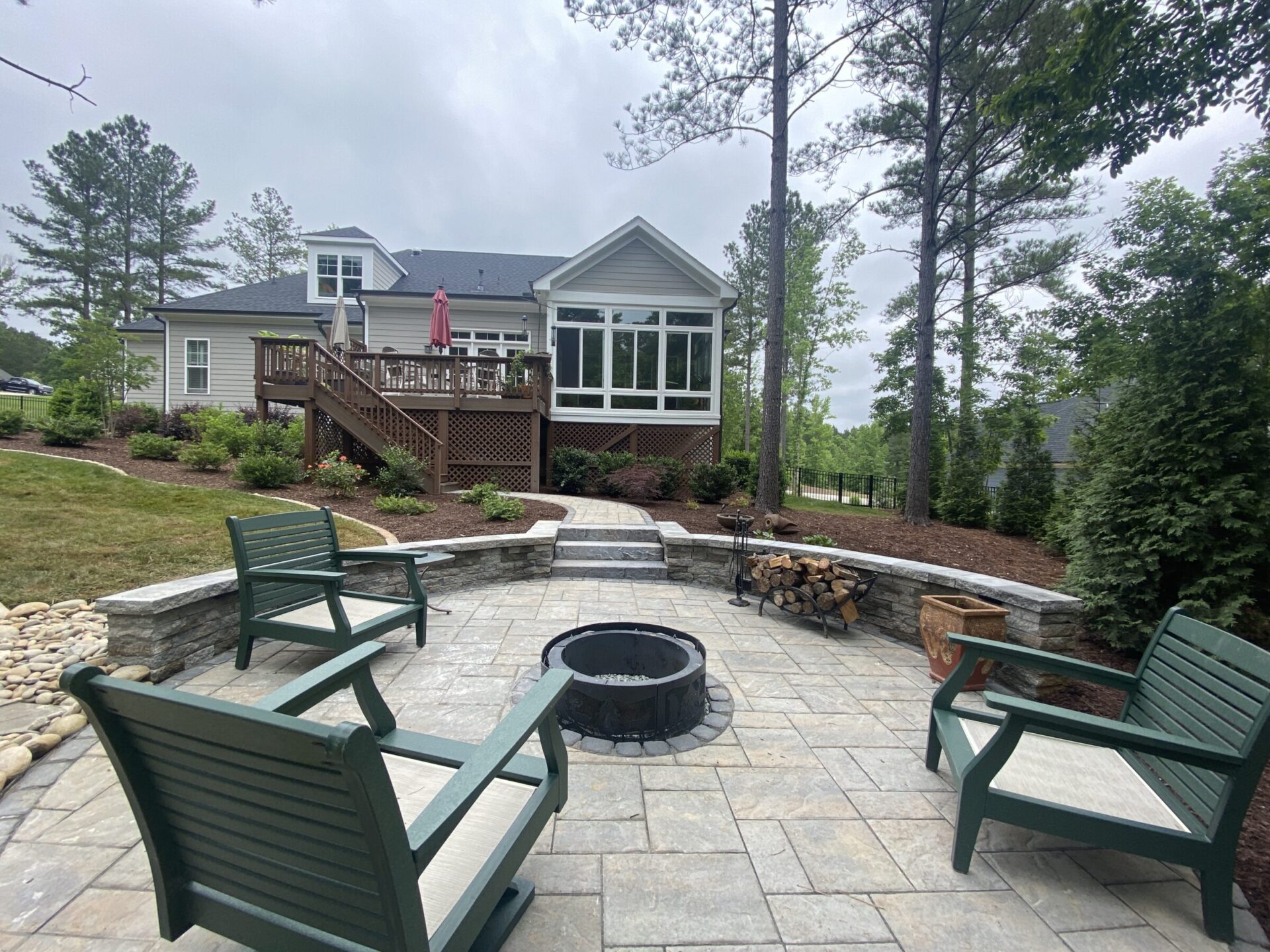 A modern house with a spacious backyard patio featuring chairs and a fire pit, surrounded by trees and landscaped gardens under a cloudy sky.