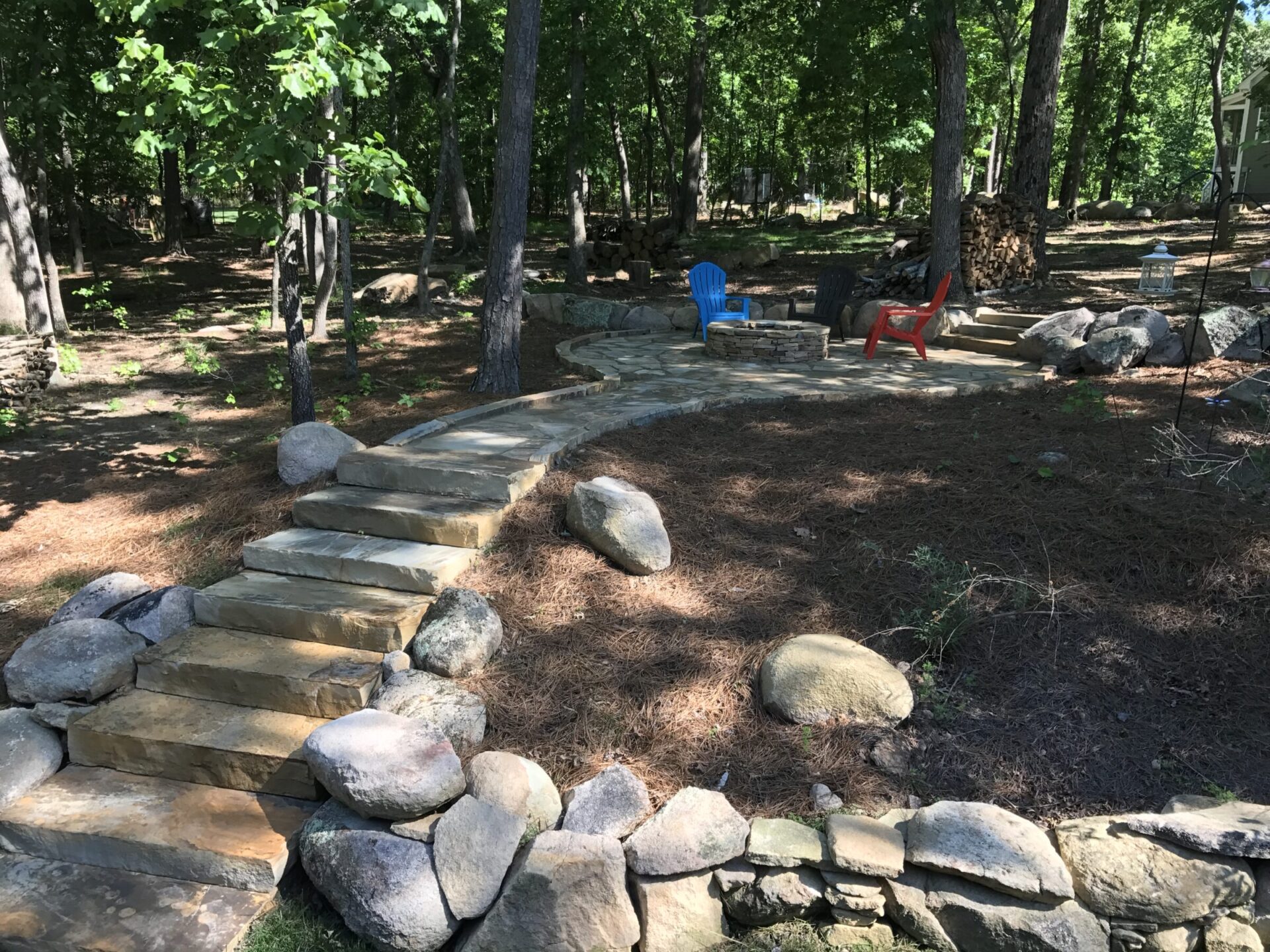 A forest setting with stone steps leads to a seating area featuring blue and red chairs, surrounded by tall trees and rocks.