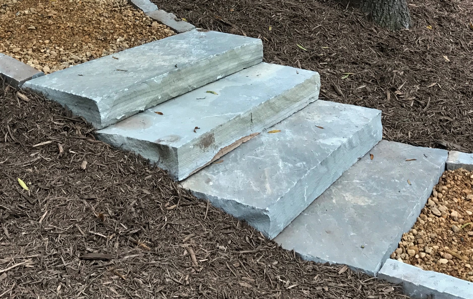 Stone steps surrounded by mulch and gravel, leading upward. Natural setting with earthy tones, possibly part of a garden or outdoor landscape.