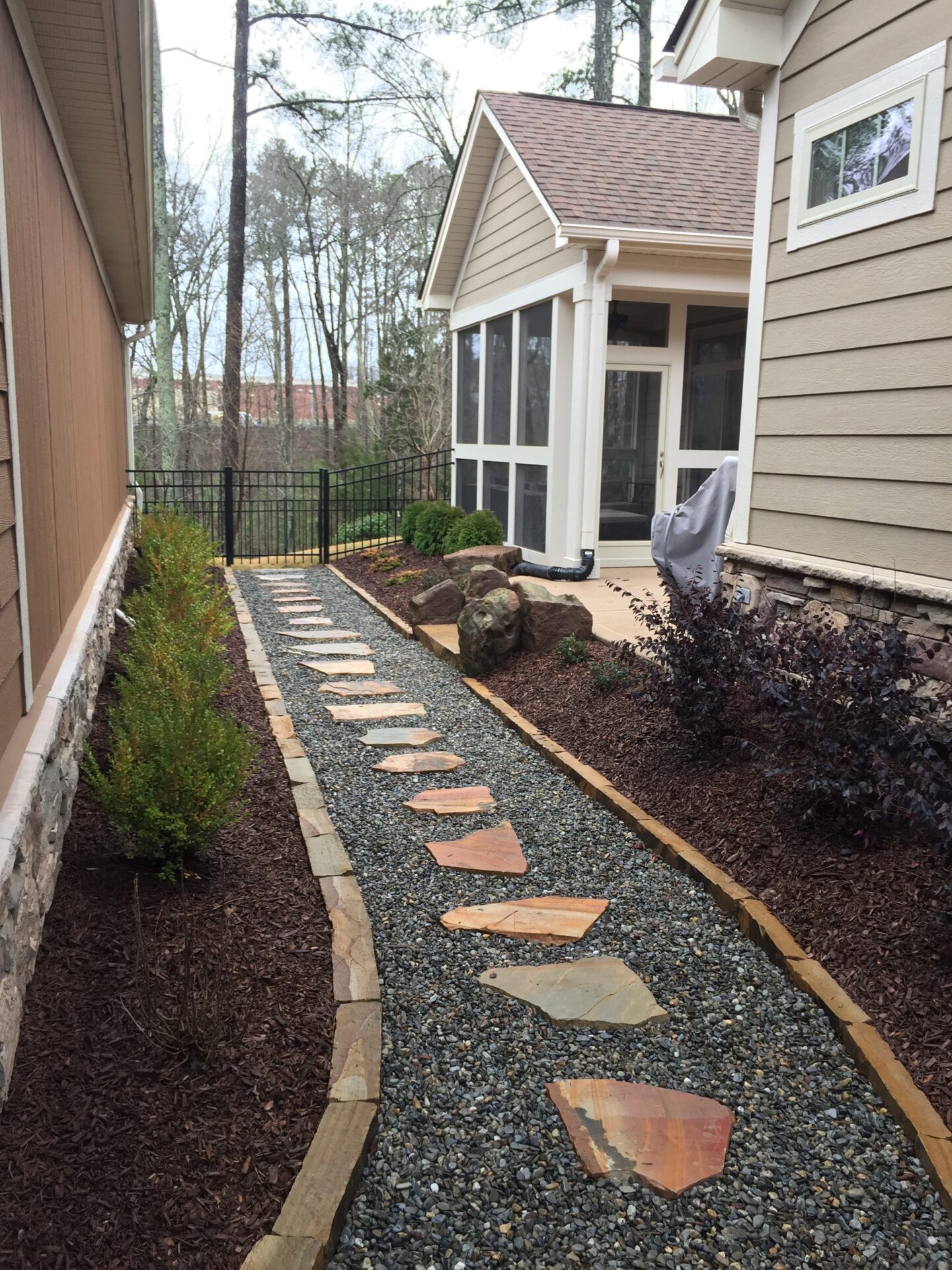 A garden pathway with stepping stones beside a house, leading to a screen porch, surrounded by mulched garden beds and small plants.