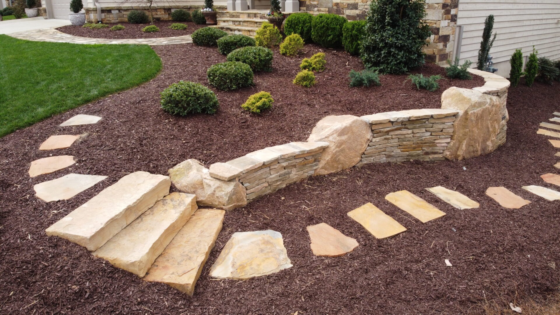 Stone steps and pathway lead through a landscaped garden with mulch and shrubs, bordered by a low stone wall, creating a serene outdoor space.