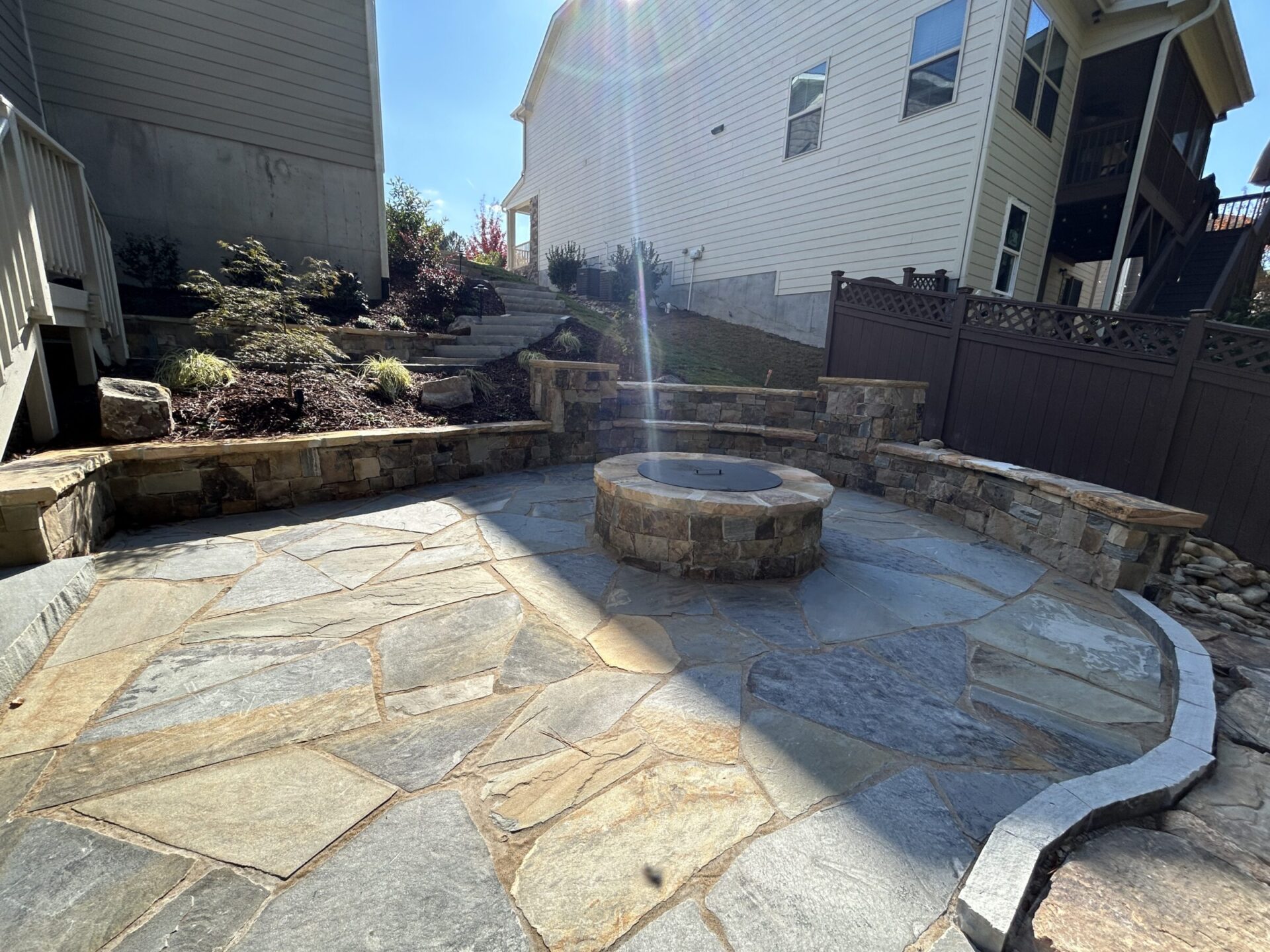 Stone patio with a circular fire pit, surrounded by landscaped garden and residential buildings, under a clear blue sky.