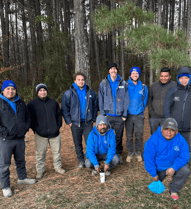 Nine persons in outdoor clothing pose in a wooded area. They wear blue hoodies and jackets, some smiling, surrounded by tall trees and pine needles.