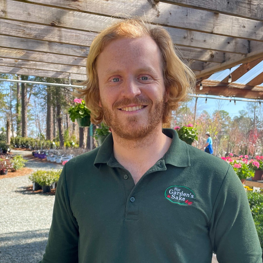 A person with red hair smiles outside in a plant nursery, wearing a green shirt with a logo. Trees and plants surround the area.