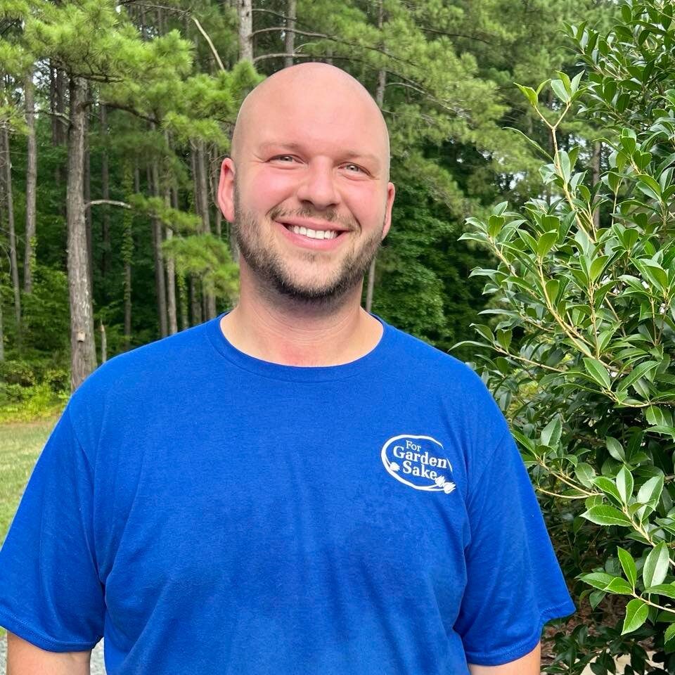 A person in a blue shirt stands outdoors, smiling, with trees and greenery in the background. No recognizable landmarks or historical buildings are visible.