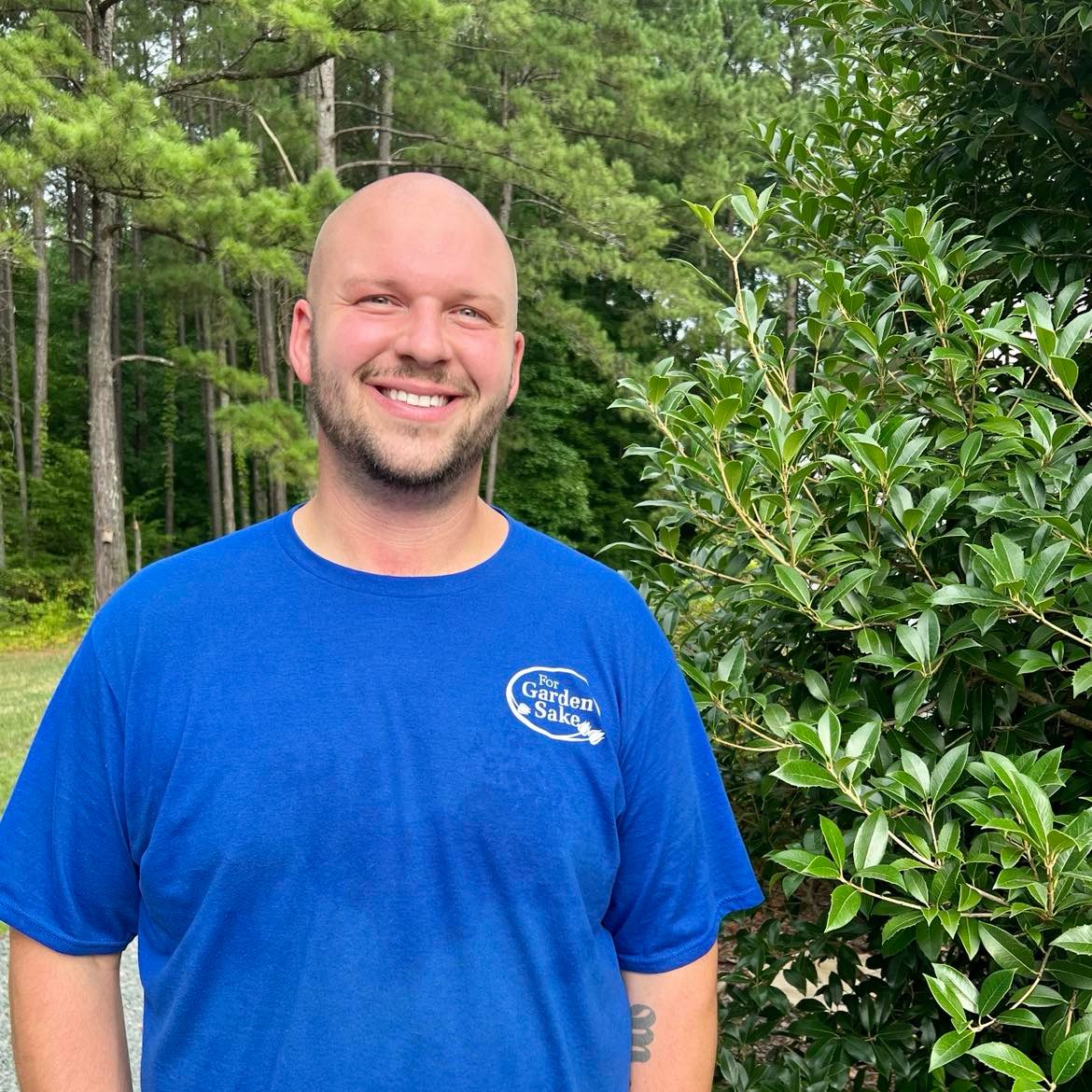 A person in a blue shirt stands outdoors, smiling, with trees and greenery in the background. No recognizable landmarks or historical buildings are visible.