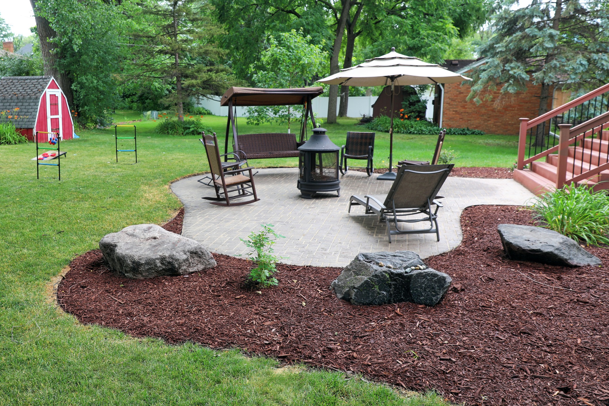 A scenic backyard features a patio with chairs, a swing, umbrella, fire pit, mulch landscaping, and green lawn surrounded by trees and a red shed.
