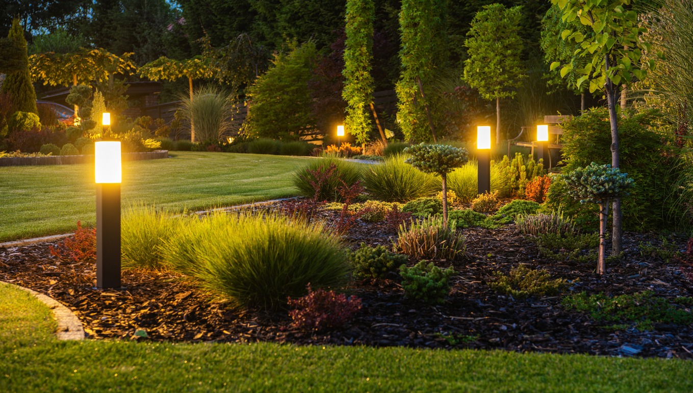 A beautifully landscaped garden is softly illuminated by a series of glowing pathway lights, surrounded by lush greenery and manicured plants at dusk.