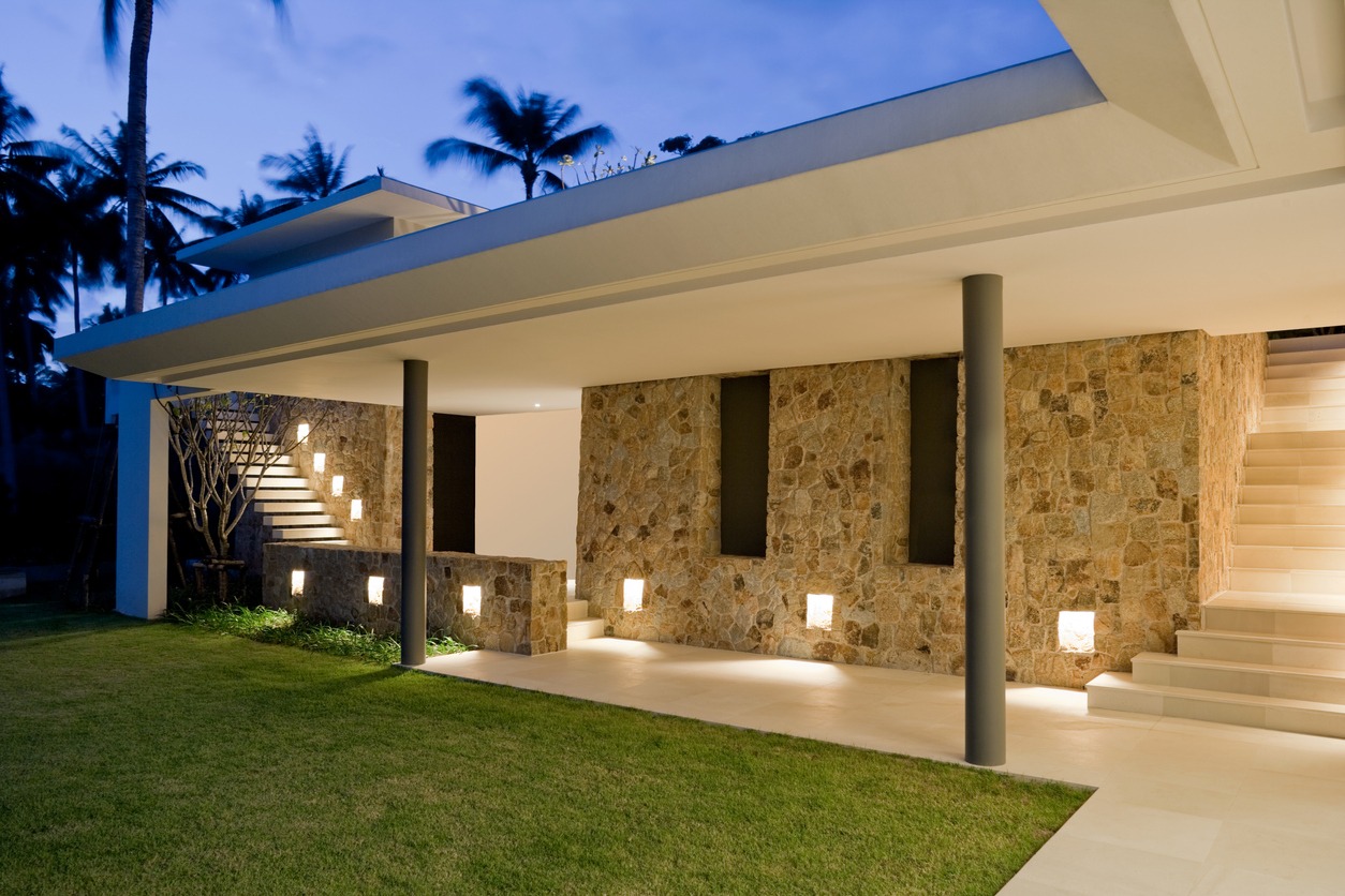 Modern stone and stucco house with outdoor lighting, stairway, and lush lawn, set against a backdrop of twilight and silhouetted palm trees.