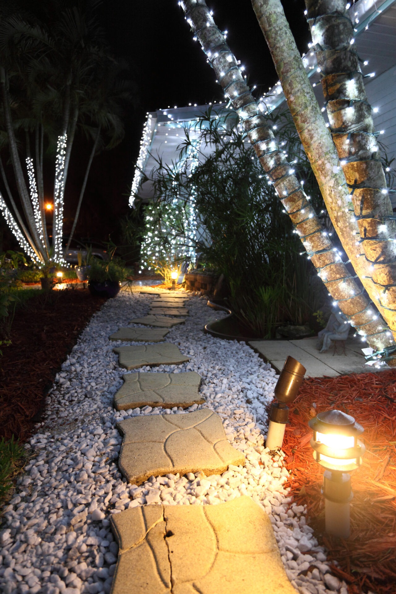 A nighttime garden scene shows a stone pathway, illuminated by string lights on palm trees and warm ground lighting, creating a cozy ambiance.