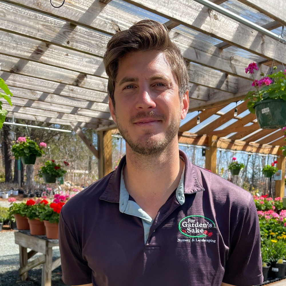 A person stands in a greenhouse surrounded by colorful flowers and plants. Sunlight filters through the wooden lattice overhead.