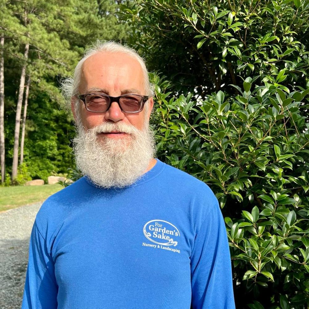 A person with a white beard and sunglasses stands outdoors, wearing a blue shirt. Green foliage and a gravel path are visible.