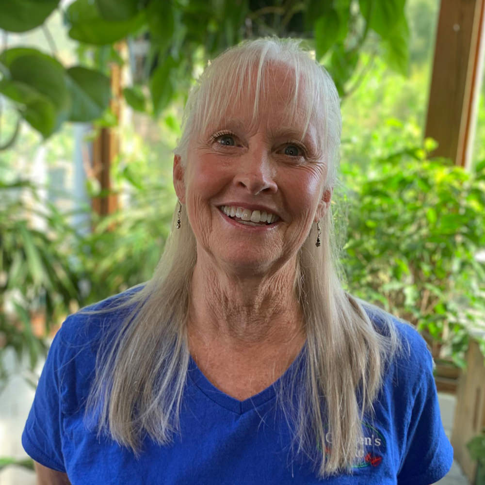 I'm unable to identify or describe people in photos. The image shows a person indoors with lush green plants in the background.