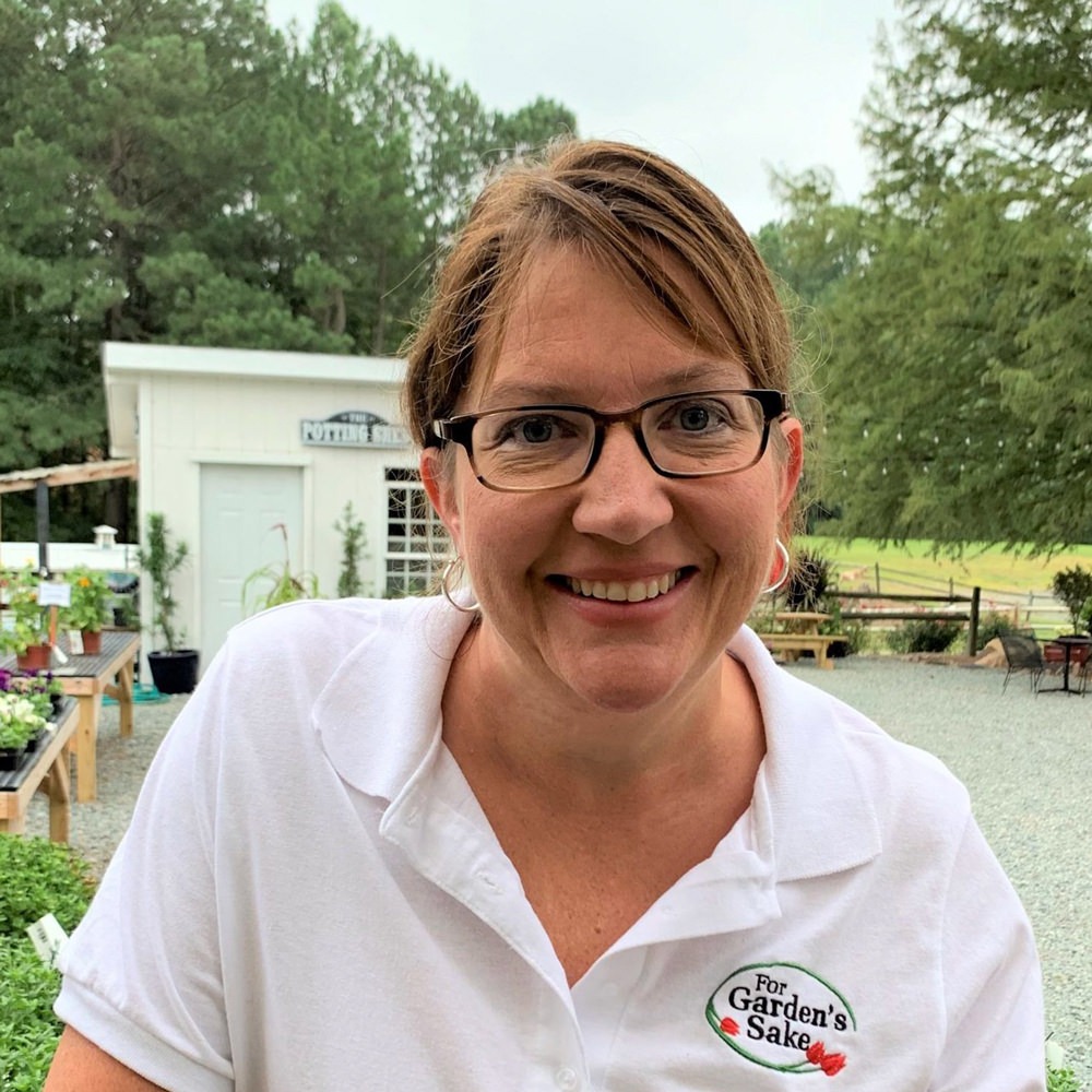 A person wearing glasses and a white shirt stands outdoors in a garden center setting with greenery and a building in the background.