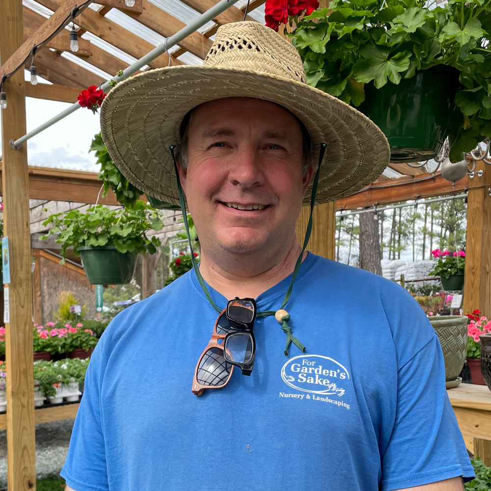 A person in a straw hat and blue shirt stands in a greenhouse, surrounded by hanging plants and flowers, smiling brightly.