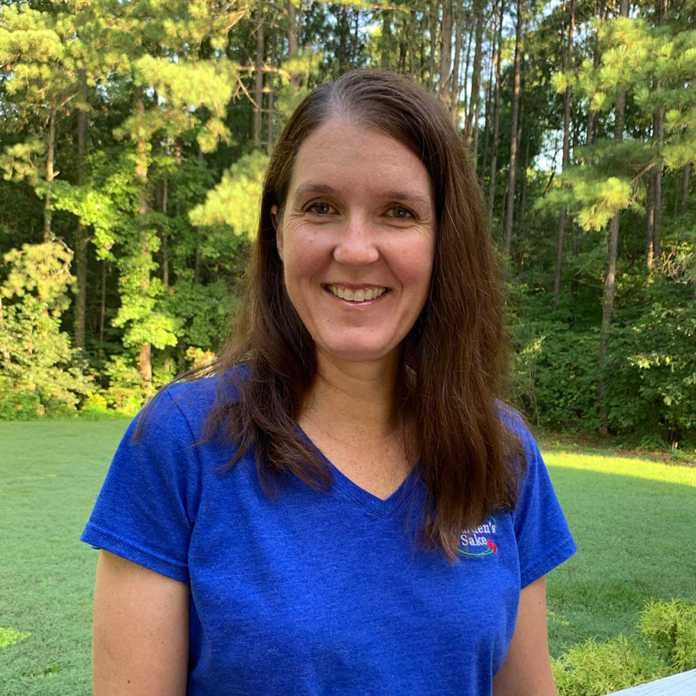 A person in a blue shirt smiles outdoors, standing in front of a lush, green forest background on a sunny day.